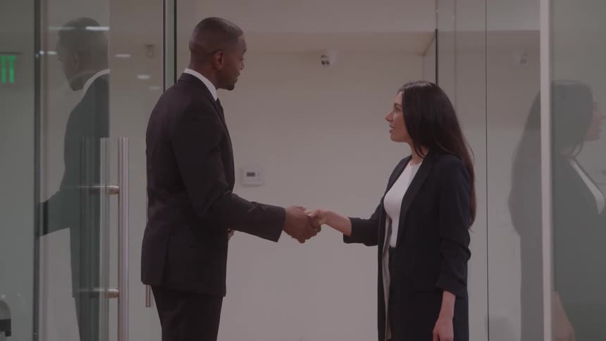 A young man and a young woman standing in an office lobby both wearing black blazers shake hands while having a discussion with glass walls behind them