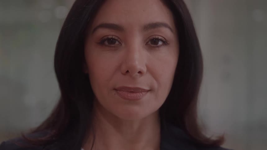 A woman with long dark hair and a serious expression wearing a dark blazer slowly transitions from having her eyes closed to looking directly into the camera with focus and intensity in a tightly framed closeup shot