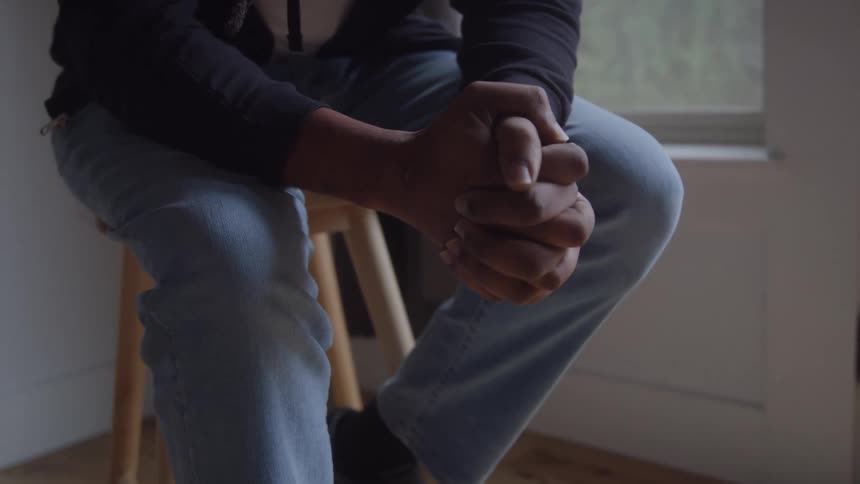 A closeup shot shows a person sitting on a wooden stool their hands clasped together with a sense of tension or contemplation the individual is wearing a dark top and lightwash jeans situated near a window that provides dim diffused light