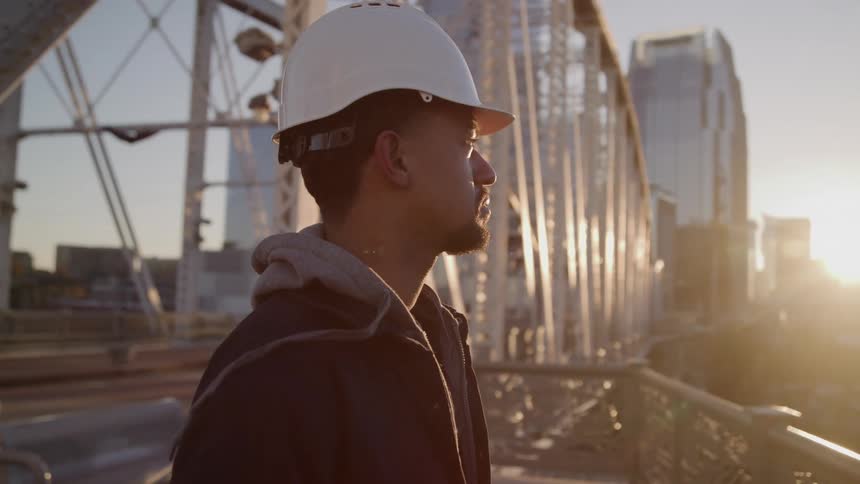 A construction worker gazes thoughtfully at a bridge during sunset