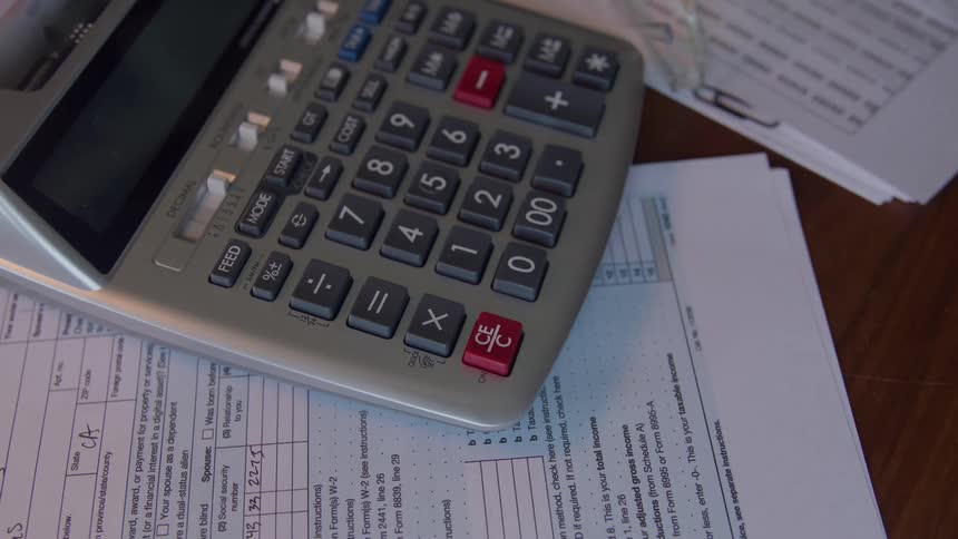 A persons hand is shown using a calculator on a wooden surface cluttered with documents