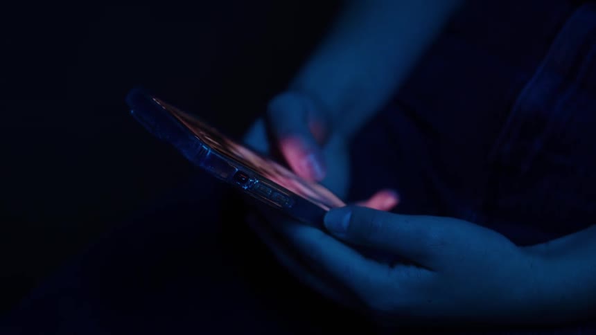 A closeup shows a persons hands bathed in blue light holding and scrolling on a smartphone in the dark