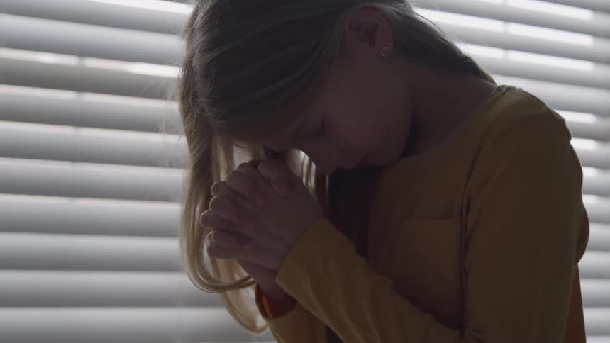A little girl is in a room with closed blinds clasping her hands in prayer with her head down and eyes closed