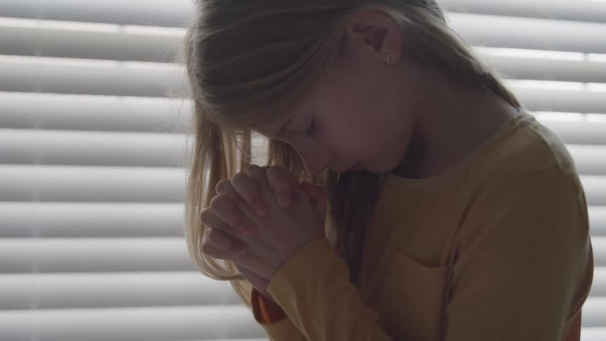A young girl is praying with her eyes closed next to a window