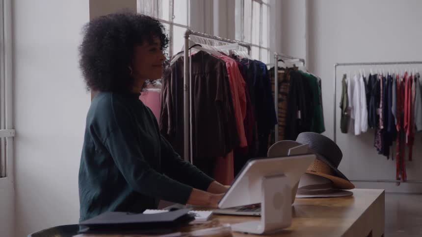 A woman with curly hair is standing behind a counter in a brightly lit clothing boutique working on a tabletstyle pointofsale system racks of clothing and large windows are visible in the background