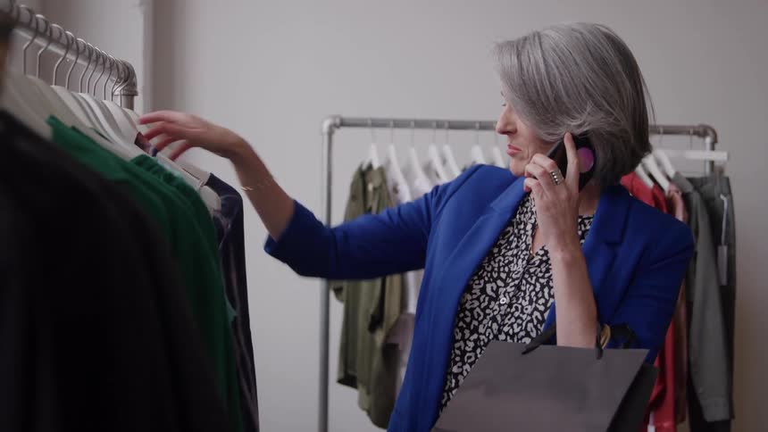 A stylish older woman with silver hair talks on her phone while browsing a rack of clothes in a boutique
