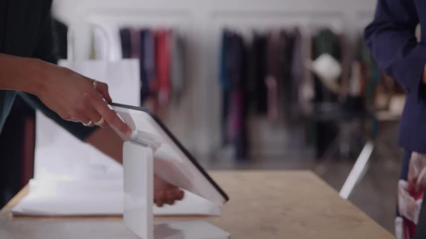 Two people stand at a check out counter in a clothing boutique the customer swipes their credit card and takes their bag