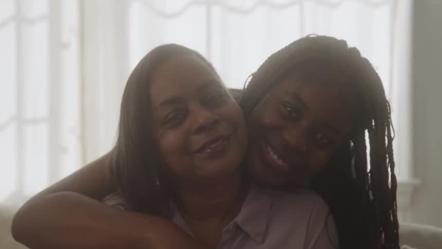 A young black female is behind an older black female hugging her as they both smile