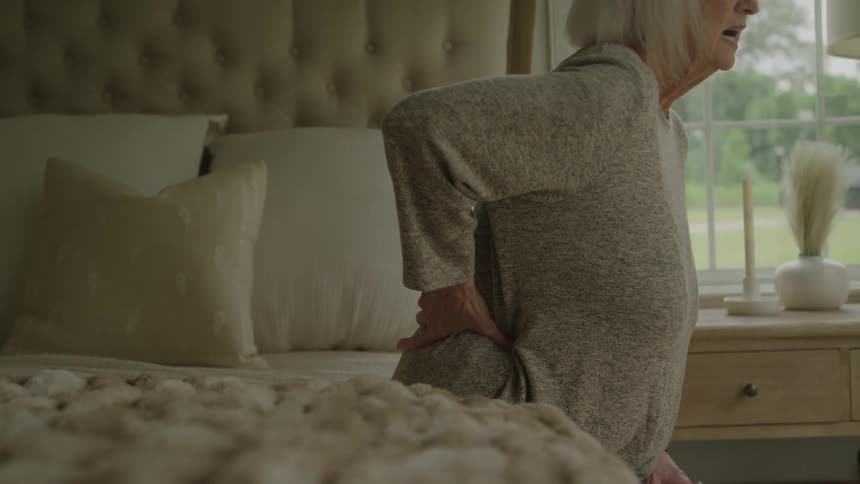 An elderly female raises from a bed grabbing her back as if in pain as she stands