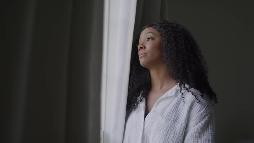 A woman looks really sad as she looks through the curtains on a window