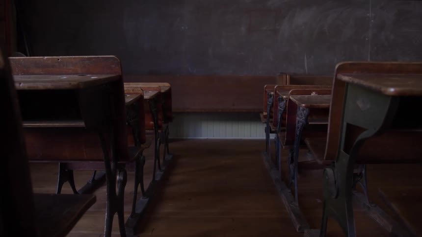 An aisle separates rows of wooden desks in an empty dimly lit vintage classroom
