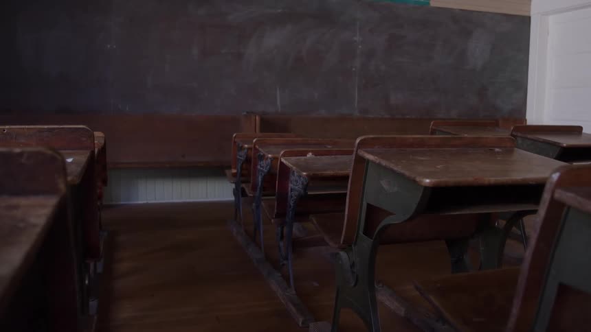 The inside of a school that looks older with a full wall chalkboard and wooden desks with a wood floor