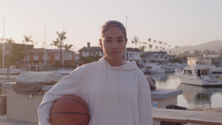 A young woman stands near a marina holding a basketball under her arm