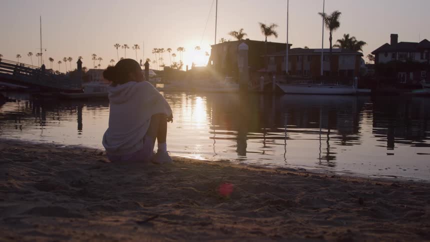 A young woman rests sitting down on the sand by a body of water near a pier with a docked boat