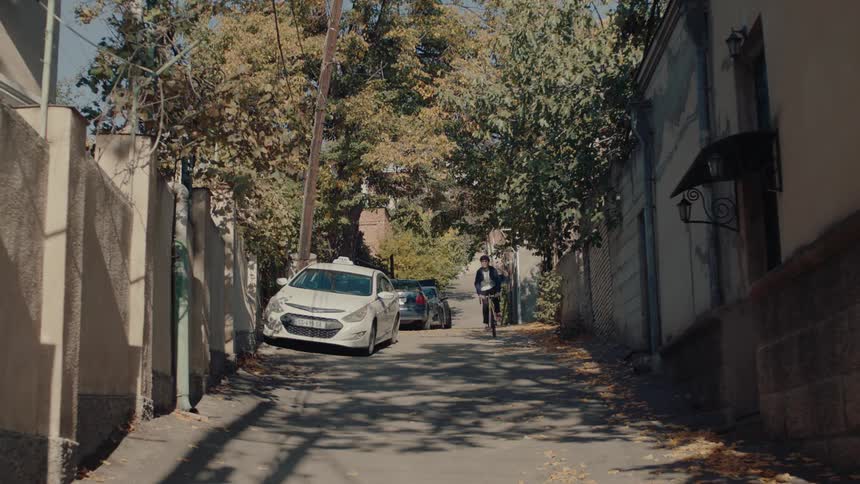 The video shows a man riding a yellow bicycle down a narrow sundappled alleyway lined with walls trees and parked cars he is wearing a dark jacket and a lightcolored hat or helmet
