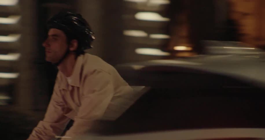 The video clip shows a man cycling on a busy city street at night weaving through traffic that includes an ambulance and several cars he is wearing a lightcolored jacket and a helmet as he rides and the bright headlights of the surrounding vehicles ar
