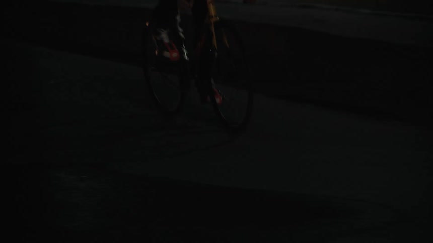 A person wearing a light blue hoodie black helmet and black pants is riding a yellow bicycle at night they are cycling through what appears to be a tunnel or underpass with concrete walls covered in faded graffiti