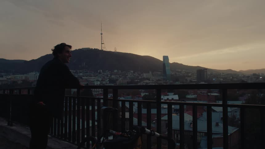 A silhouetted man stands next to his bicycle on a bridge at dawn looking out over a city and a distant mountain