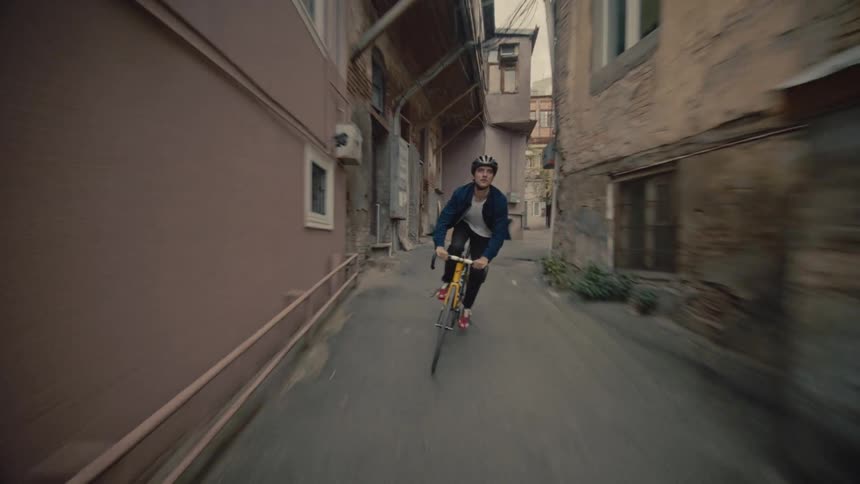 A cyclist wearing a helmet rides quickly through a narrow alleyway lined with old buildings and muted colors