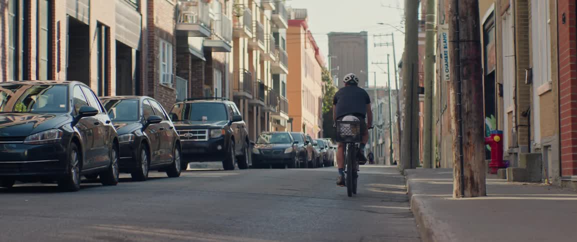 A cyclist viewed from the rear and wearing a helmet is riding away from the camera down the middle of a narrow sunlit urban street lined on both sides by parked cars and brick apartment buildings