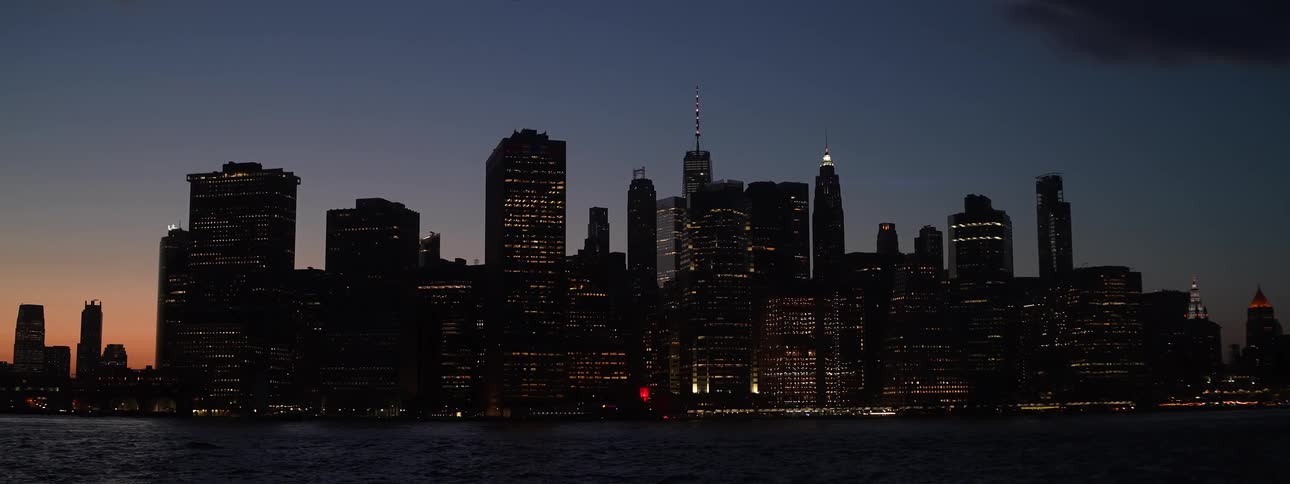 A city skyline is silhouetted against the twilight sky as its lights begin to illuminate the buildings all viewed from across the water