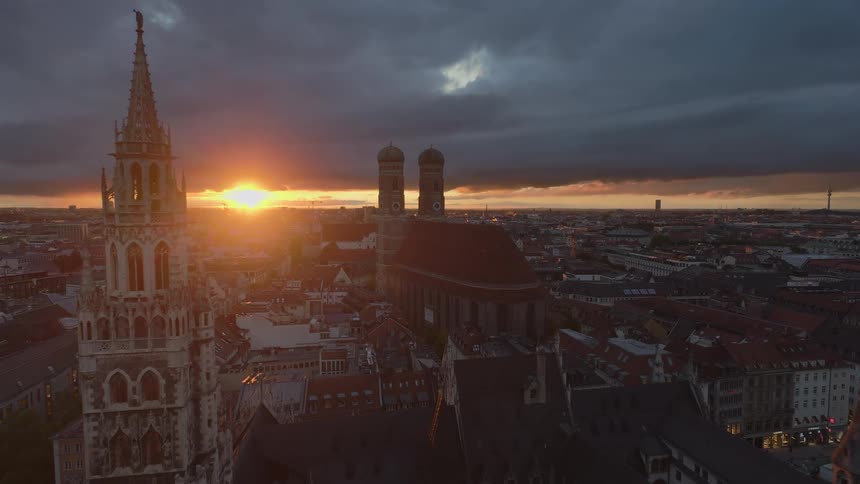 An aerial drone shot begins high above a densely packed city at dusk revealing the bright orange sunset on the horizon and the illuminated towers across the cityscape