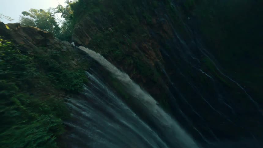 A drone shot along the massive vertical cliff face of a waterfall draped in vibrant green tropical foliage capturing the water cascading down in multiple streams illuminated by bright sun