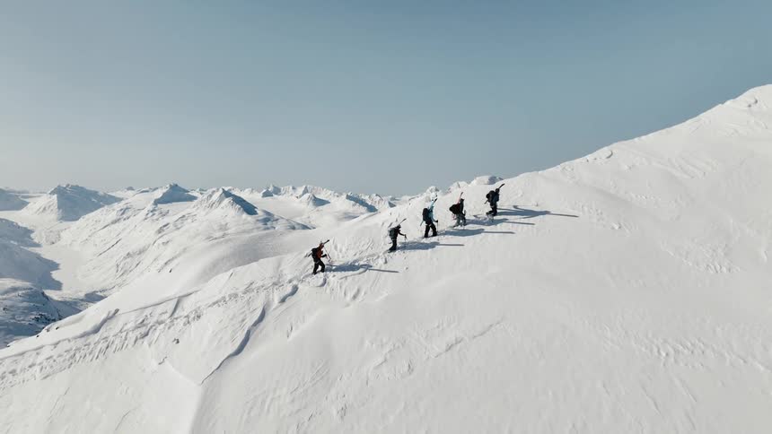 An aerial shot flies over a group of five trekkers as they hike in single file up a steep snowy mountain ridge revealing the endless sunlit peaks beyond
