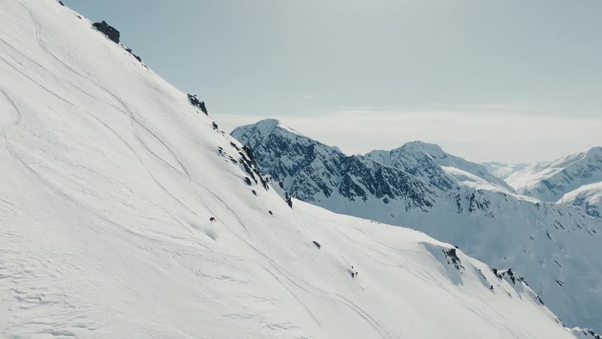 An aerial view follows a skier in a red jacket as they descend a steep wideopen mountain face carving turns through deep snow under a bright hazy sky