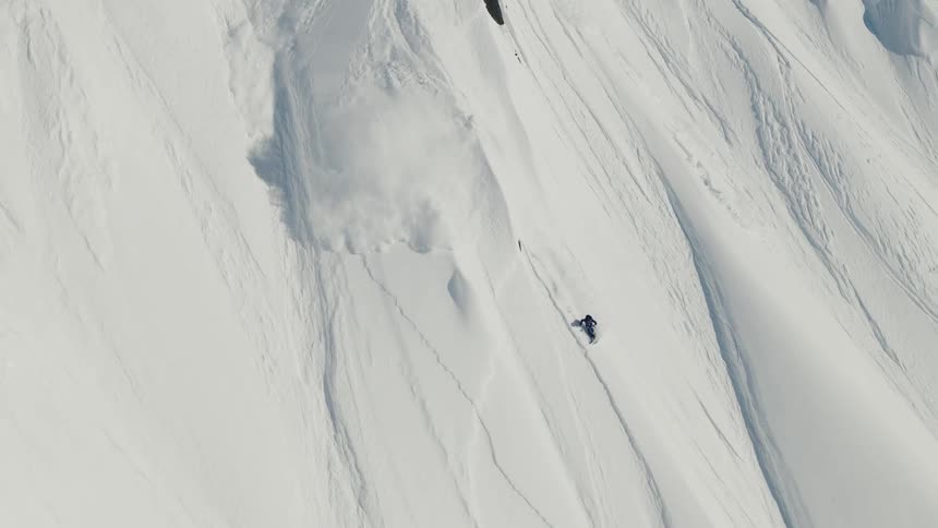 Several climbers and skiers traverse a steep snowcovered mountain ridge under clear blue skies with a few descending downhill and a distant sea visible beyond the snowy peaks