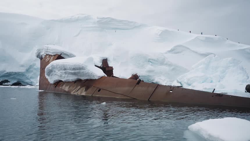 A rusted shipwreck partially buried in snow and ice rests in calm polar waters with a few people visible trekking along the snowy ridge in the background