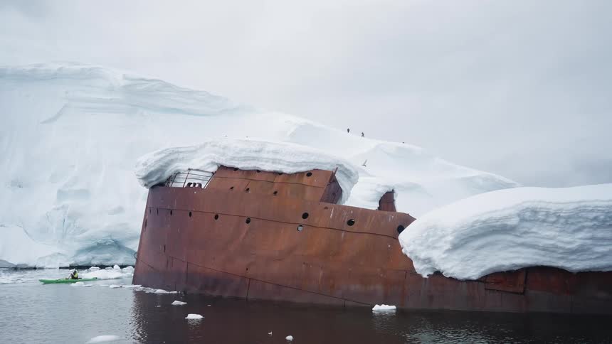 Man in a canoe sails by an old snow covered shipwreck along the arctic coast as birds fly overhead
