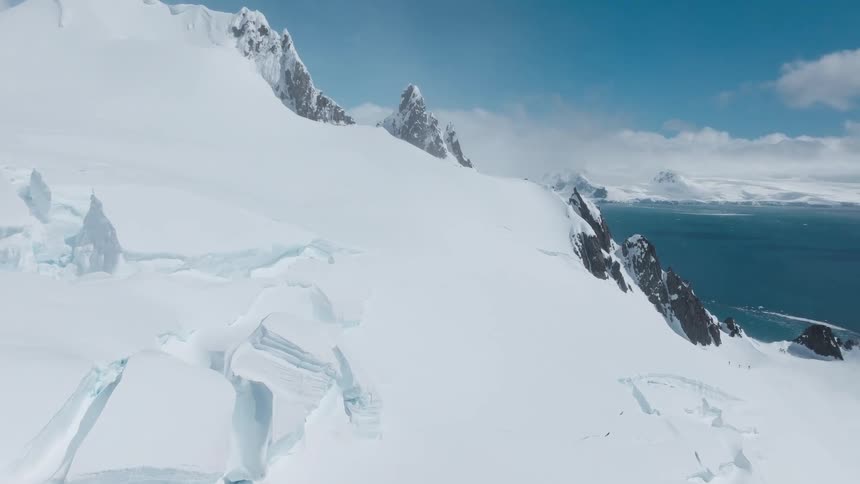 Snowcovered mountain range ends at a blue sea under a partly cloudy sky