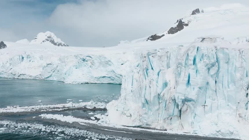 Towering bluewhite glacier cliffs rise above the icy shoreline their massive faces streaked with cracks and melt lines under a pale sky