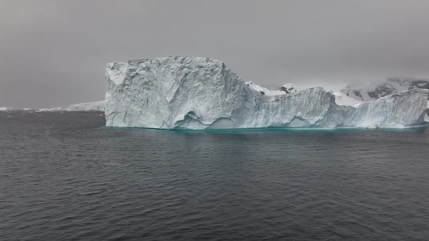 A massive iceberg with a jagged surface floats in dark cold ocean waters against a backdrop of snowy mountains