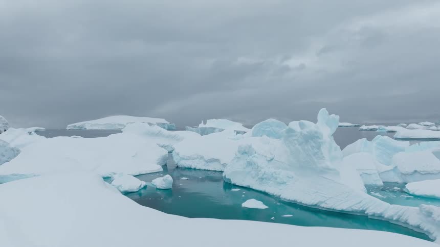 An aerial view flies over a field of massive icebergs floating in deep bluegreen water beneath a heavy overcast grey sky