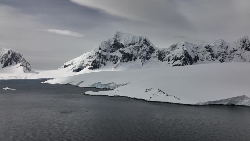 An aerial view slowly drifts across a vast icy landscape revealing jagged snowcovered mountains rising dramatically from the dark calm water of an arctic bay
