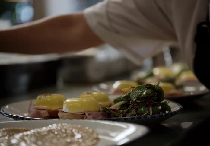 A chef uses a large metal ladle to pour rich yellow hollandaise sauce over several eggs benedict dishes lined up on a counter the finished plates which also include side salads and toasted bread are then prepared for service