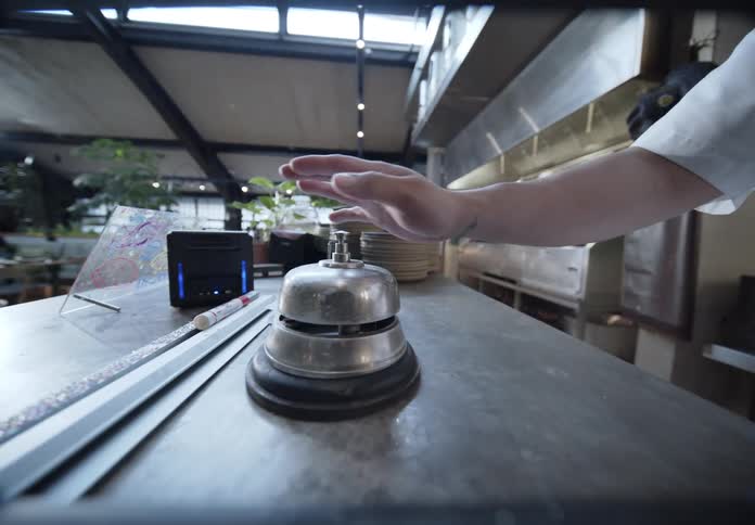 In a restaurant a chefs hand reaches across the kitchen pass to ring a silver service bell