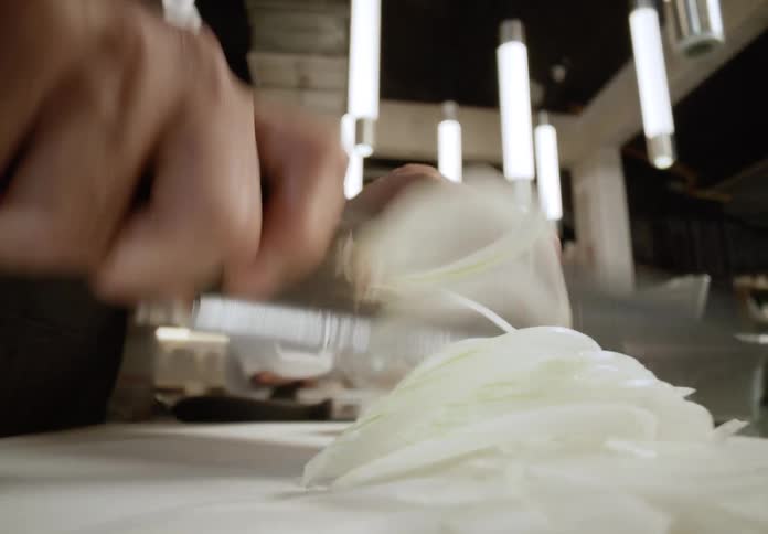 The hands of a chef dressed in a white uniform and a dark apron slicing a white onion into thin strips on a cutting board with a large knife while cylindrical pendant lights hang from the dark ceiling of the professional kitchen setting