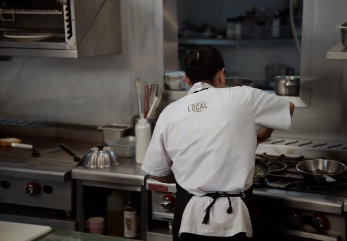 Two female chefs wearing white uniforms and black aprons are working in a commercial kitchen they briefly turn to talk to each other and smile before one turns back to a stove to attend to a pan