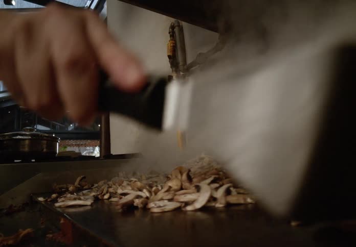 A cook in a restaurant kitchen prepares steaming sliced mushrooms on a hot grill