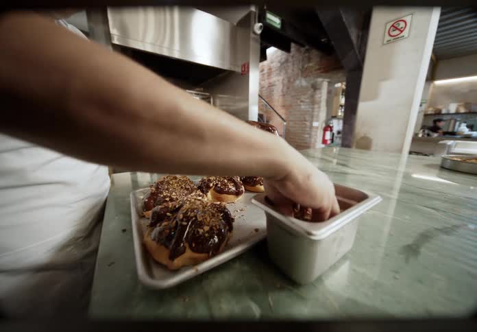 A baker in a restaurant is drizzling chocolate and nuts on pastries