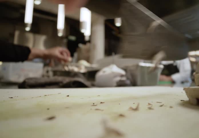 Closeup shot focuses on the repetitive motion of a textured chefs knife as a cooks hands quickly slice white mushrooms on a cutting board with the blurred background showing a dimly lit professional kitchen with another chef looking