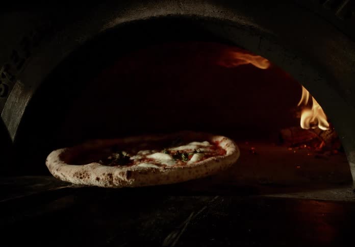 A freshly baked pizza is pulled out of a dark wood fired oven with flames glowing in the background