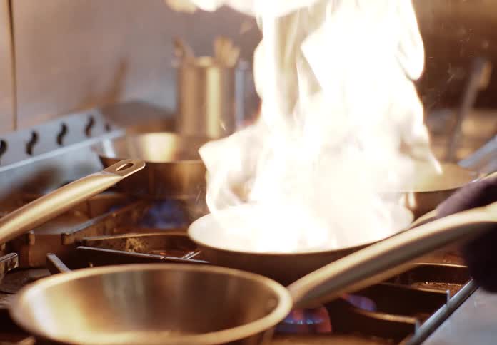 Chef squirts fluid into a skillet and jiggles the burning skillet as the flames rise high