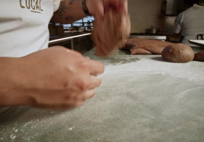 A baker sprinkles flour onto a countertop and expertly kneads a loaf of brown dough
