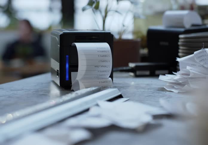 Two receipt printers on a counter in a well lit business are printing receipts