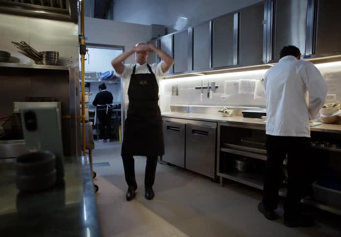 The man wearing the apron is in a kitchen making some dance gestures while someone next to him is cooking