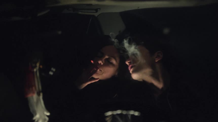 Viewed through the windshield a man and woman huddle in a dark car as she smokes a cigarette the ember glowing brightly before smoke fills the air